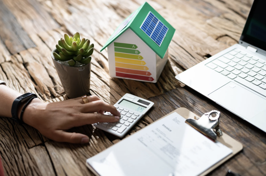 Main sur une calculatrice avec une maquette de maison et une étiquette énergétique – analyse financière du logement social et rénovation thermique.