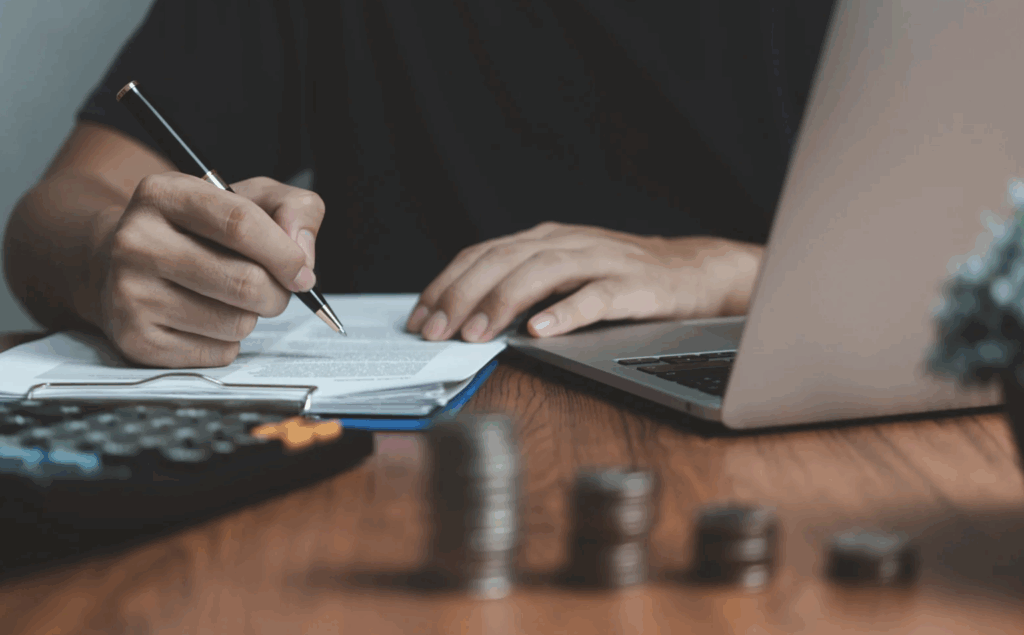 Personne écrivant sur un document posé sur un bureau, avec un ordinateur portable, une calculatrice et des piles de pièces.