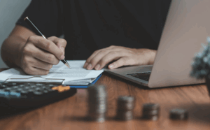 Personne écrivant sur un document posé sur un bureau, avec un ordinateur portable, une calculatrice et des piles de pièces.